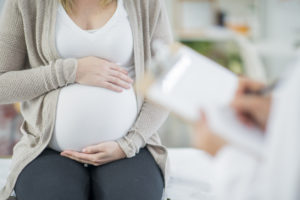 A pregnant Caucasian woman is indoors in a doctor's office. Her female doctor is wearing medical clothing. The woman is holding her stomach which the doctor writes on a clipboard.