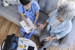 In this bird's eye view, a female doctor sits on a living room couch with a senior female patient. She holds a clipboard as the patient gestures and speaks. There is a doctor's bag and medical equipment on the coffee table.