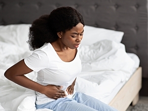 Young Black woman sitting on edge of bed holding abdomen in pain, pelvic pain endometriosis concept