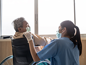 Elderly man in wheelchair in front of hospital windows, with female nurse wearing mask and gloves
