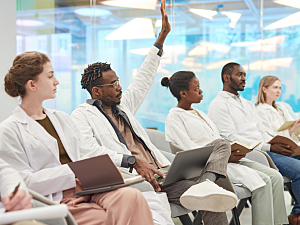 Diverse group of men and women in white lab coats learning, arm raised to ask question