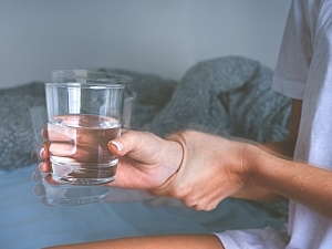 Woman sitting on bed holding glass with shaking hand, suffering from essential tremor
