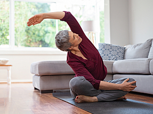 Older woman praciticing yoga in living room, sitting on mat stretching
