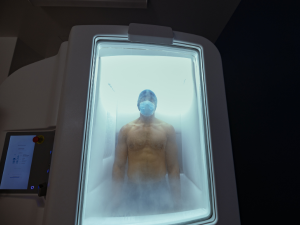 A person wearing a mask stands inside a cryotherapy chamber filled with mist, with part of a control panel visible to the side.